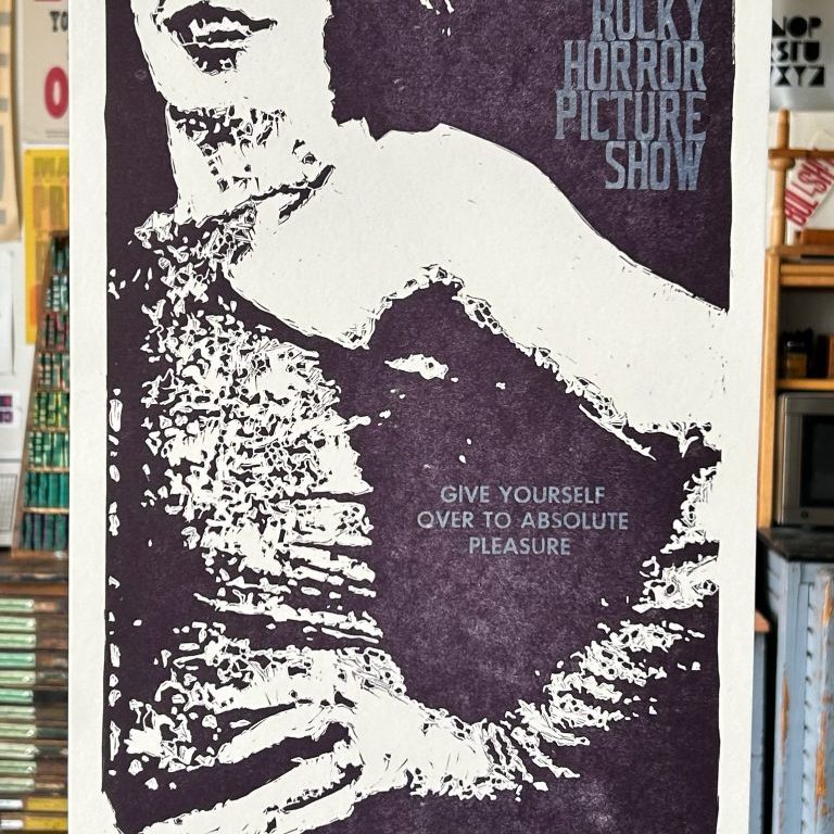 A picture of a letterpress printed poster hanging in a print shop. The print is a purple linoleum carving of Dr. Frank n Furter from the Rocky Horror Picture Show. The text in the top right corner reads Rocky Horror Picture Show with the center text reading give yourself over to absolute pleasure.