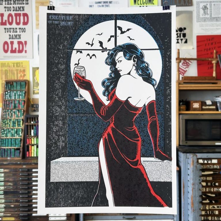 A letterpress printed poster hangs in front of a print shop featuring an comic-style illustration of a vampire looking over her shoulder wearing a red satin dress holding a wine glass in her hand. She stands in front of a window leaning against a ledge. Outside the window, a trail of bats fly over the moon.