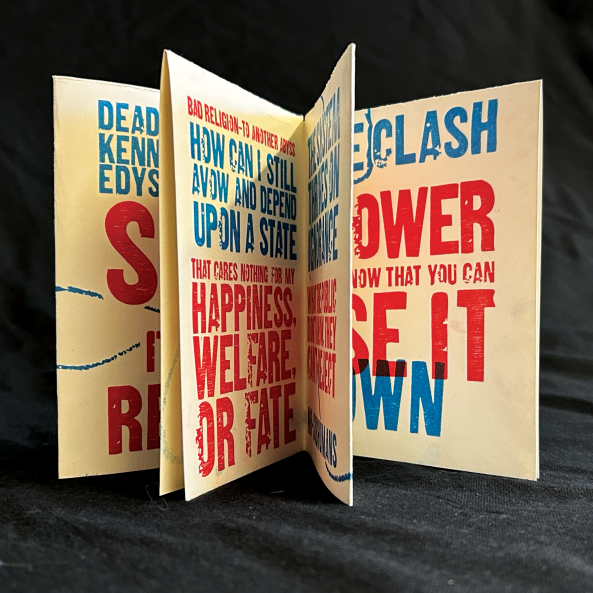 A zine stood up so that the internal pages are semi-visible in front of a black sheet background. The zine is letterpress printed with a bold sans-serif wood type with its text alternating between red and blue.
